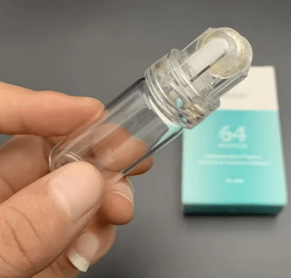 Hand holding a clear glass microneedle roller with a white and gold roller head.